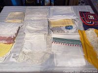 Photo showing assorted vintage linens folded and laid out on table including crochet, embroidered, lace, printed and plain fabrics in white, cream, yellow, pink and multicolor patterns.