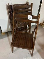 Four brown wooden folding chairs stacked and folded against a wall, showing front and side views. Chairs have slatted seats and backs, used condition with scratches and wear visible.