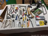 Photo showing a wide range of hand tools laid out on a white surface, including clamps, pliers, hammers, wrenches, screwdrivers, measuring tape, hand saw, and files.