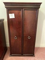 Front view of closed media cabinet showing dark wood finish, decorative molding, and metal ring pull handles on two doors.