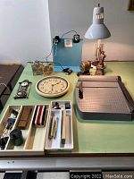Photo showing various office items including paper cutter, clock, metal file divider, desk lamp, pencil sharpener, Statue of Liberty decor and small office supplies arranged on a table.