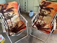 Two outdoor folding chairs with white metal frames and orange fabric seats and backs featuring palm tree sunset print, placed on a concrete floor in storage.