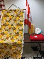 Full view of the hammock fabric hanging on a metal chain with a floral pattern and fringe, folded red blanket, sun hats, umbrella and Canadian flag on table in background