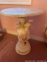 Full view of Roman style pedestal table with cream color, round top, and sculpted base of three helmeted heads