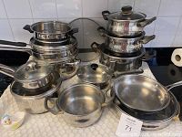 Full view of stacked stainless steel pots and pans with glass lids on a stove, showing varying sizes and some black handles.
