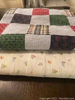 Two folded quilts stacked, top quilt has multi-colored patchwork squares, bottom is cream with small colorful sailboat motifs.