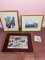Three framed art prints displayed together on carpet with light pink wall background, showing varied urban and water scenes.
