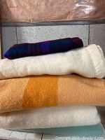 Stack of four assorted wool blankets including a tan and orange striped Hudson's Bay Point Blanket, cream-white blanket with satin edging, dark blue/maroon plaid blanket, and a pale blanket underneath.