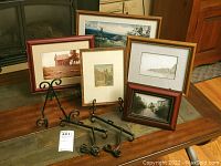 Five framed prints arranged on a wooden surface with seven wrought-iron easels in front