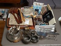 Overall view of the items on a table showing wooden servers, glass bowl, pewter and silverplate pieces, serving boards, spoons and other utensils.