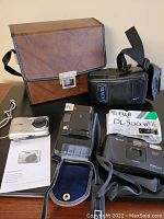 Full lot view showing binoculars case, Kodak digital camera, Minolta and Fuji cameras, and accessories arranged on a flat surface