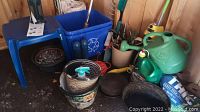 Photo of assorted garden tools, watering cans, plastic pots, and blue recycling bin on shed floor.