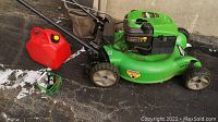 Full view of green Lawn-Boy mower with red fuel can and small green basket holding key and charger on asphalt surface.