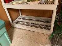 White plastic shelving unit with two slatted shelves, visible bottom shelf holding household items, placed on tiled floor.