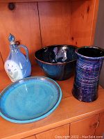 Four pottery items displayed together on a wood surface: blue jug with goat design, large dark blue bowl, purple ribbed vase, and turquoise blue plate.