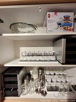 Wide view of cupboard containing glass spice bottles with white lids, spice jars, salt and pepper shakers, metal containers, and two black storage racks.