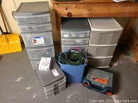 Three rolling storage units with clear/gray drawers and a blue waste bin containing yellow and green extension cords, plus a gray carry case on concrete floor.