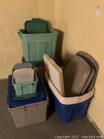 Five large and three smaller plastic storage totes stacked and arranged with multiple lids in a corner.