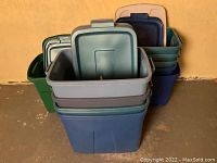 Stack of various large plastic storage totes including Rubbermaid and Sterilite in blue, green, and gray shades with lids on top.