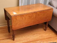 Photo shows vintage dropleaf table in closed position with drop leaf on one side, revealing warm wood tone and surface wear marks.