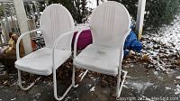 Pair of white vintage metal chairs outdoors, showing rounded backs and worn paint finish.