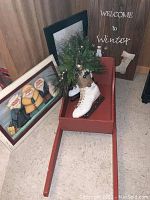 Full view of wooden red sleigh containing white vintage-style ice skate with greenery decoration, with folk art Santa prints and winter signs in background