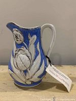Front and side view of antique porcelain pitcher showing blue crackled glaze and raised white floral reliefs with silver accents.