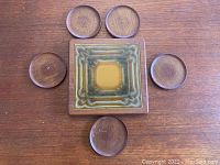 Top-down view of a square Danish teak trivet with a glazed ceramic insert in earth tones, surrounded by five round teak coasters