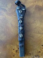 Full view of the carved wooden object showing its overall shape, embedded cowrie shells, and leather binding on a wooden surface.