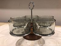 View of five clear glass condiment containers with perforated metal bases fitted into metal frame on a circular teak base.