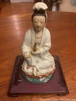 Porcelain figurine of Kwan Yin seated on a wooden stand, front view showing facial features and detail of robe and hands.
