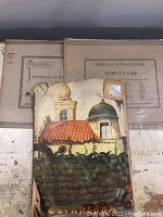 Two closed beige portfolios resting against a wall, one labeled 'Portfolio of Photographs World's Fair'. A smaller painted cover scrapbook is in front.