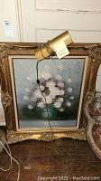Full view of floral oil painting in gold ornate frame with lighting attached at top