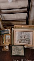 Blanket stand with nine framed artworks leaning against a wall, showing a variety of frame styles and art themes including landscapes and street scenes.