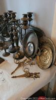 Photo of two large dark patina metal multi-arm candelabras with three or more arms, decorative bases, and several smaller single candle holders close by. Various serving trays and decorative metal items in background.