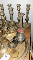 Multiple tall and medium sized brass candlestick holders with ornate floral and decorative bases arranged on a wooden round table with additional small metal items.