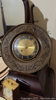 Ornate round wall clock with decorated border and brass face, on a wooden surface with part of a mirror in background.