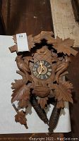 Full view of the wood carved cuckoo clock with two bird carvings, roman numeral clock face, two pine cone shaped weights, and leaf carvings.