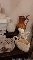 Display of white Greek head busts, white ceramic vases and pots, light brown ornamental ceramic pitcher, and ceramic basket with flower