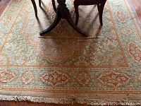Area rug beneath dining table showing intricate traditional design in beige, teal and rust orange colors with fringed edges.