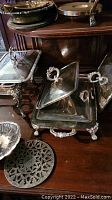 Photo showing multiple silver plated serving pieces including covered chafing dishes with ornate handles and legs placed on wooden surfaces.
