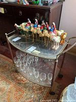 Photo of the tea cart showing camel figurines on top glass shelf and clear glasses on bottom shelf, taken to display the contents and condition.