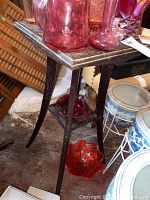 Photo showing close-up of tabletop and legs of one wood pedestal stand with pink glass vases on top, highlighting worn and scratched surface.