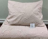 Photo showing the light pink comforter and one matching sham with a textured floral pattern, laid flat against a wall and floor.