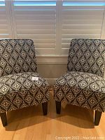 Front angle showing pair of armless accent chairs with black and white geometric upholstery on hardwood floor in front of window shutters