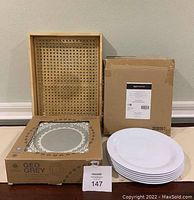 Photo showing packaging of ceramic Geo Grey plates, 4 in set, and a stack of 6 white melamine plates. Natural wood and cane serving tray behind with woven cane design.