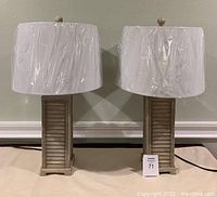 Pair of wood tone shutter-style base table lamps with white drum shades still wrapped in plastic, placed side by side.