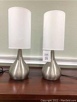 Two silver table lamps side by side on a wooden surface, both with bulbous rounded brushed silver bases and tall white cylindrical shades.