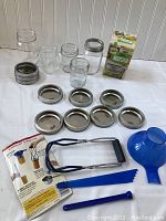 Photo showing multiple clear glass Ball canning jars, several metal lids and bands, blue plastic funnel, jar lifter tool, and canning instruction manual spread on a white table with beadboard background.