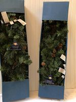 Two boxes each containing a Columbia Spruce garland with green artificial pine branches and brown pine cones, packaged in blue rectangular boxes.